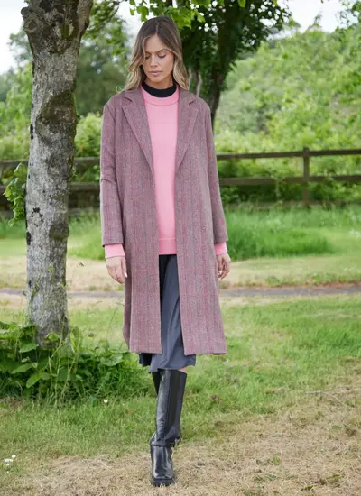 Woman wearing a brown and pink herringbone Irish tweed duster coat with lapel collar, styled over a pink sweater and black boots.